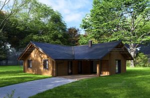 Blockbohlenhaus mit modernem Winkel-Grundriss, dunklem Dach und überdachter Terrasse im Grünen