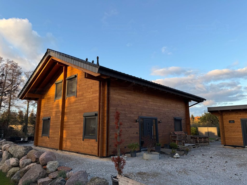 Blockbohlenhaus „Drebkau“ mit moderner Doppelblockbohlen-Konstruktion, großer Eingangstür, natürlichen Holzfassaden und einer harmonisch angelegten Umgebung. Im Hintergrund blauer Himmel und ländliche Atmosphäre.
