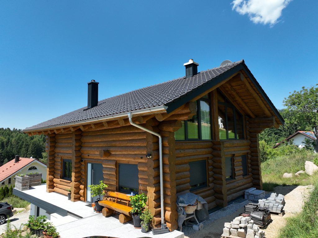 Schrägansicht eines Naturstammhauses mit grauem Satteldach, zwei Kaminen, breiten Dachüberständen und sichtbarer Rundstammkonstruktion