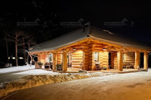 Naturstammhaus „Potsdam“ aus massiven Rundstämmen mit verschneiter Umgebung und warmer Außenbeleuchtung bei Nacht