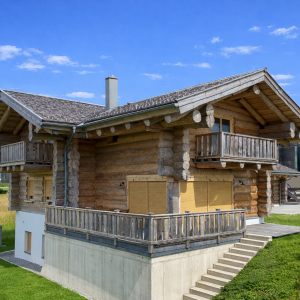 Naturstammhaus mit zwei Holzbalkonen an der Giebelseite, hohem Betonsockelgeschoss, seitlicher Außentreppe aus Betonstufen und großen Fenstern mit gelben Rollläden