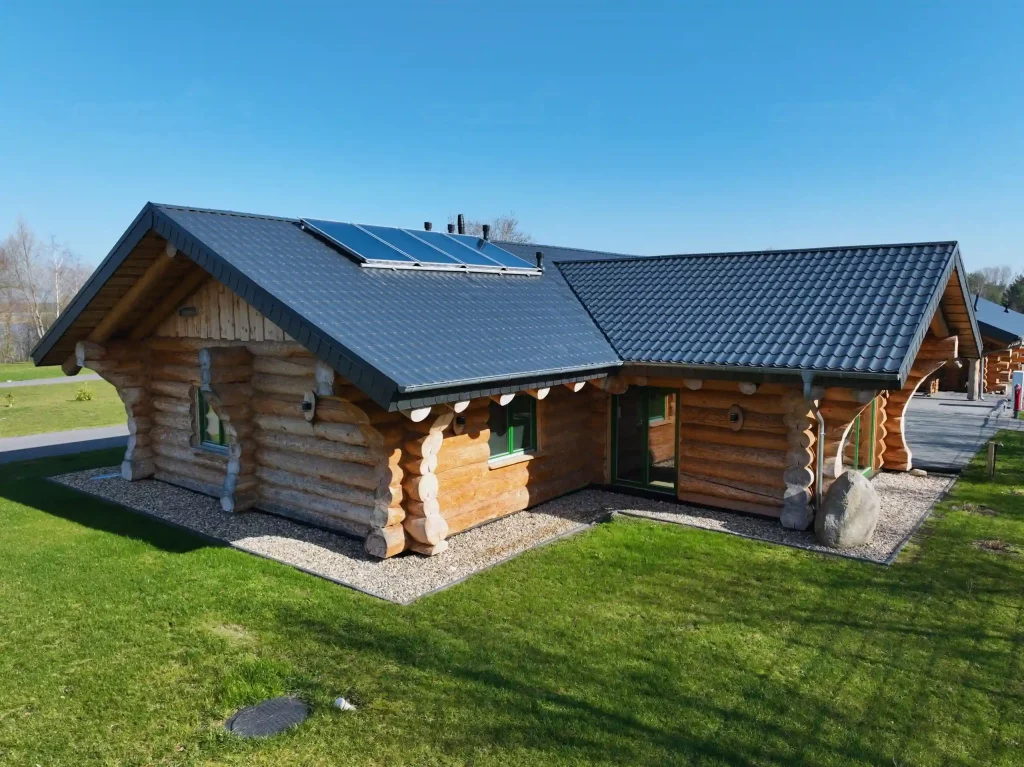 Außenansicht eines Naturstammhauses Modell Hamburg mit massivem Rundholz, dunklem Satteldach, integrierter Solaranlage und grünen Fensterrahmen auf gepflegtem Grundstück