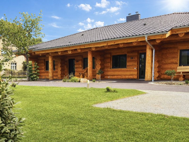 Vorderansicht des Naturstammhauses Bodensee mit überdachtem Eingangsbereich, massiven Rundholzstämmen und breitem Dachüberstand im Gartenbereich