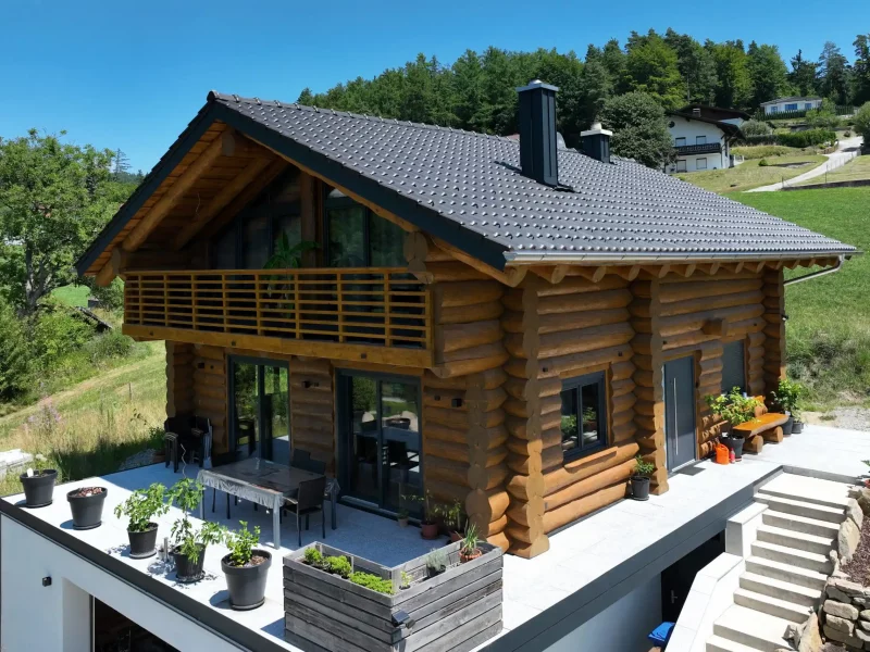 Außenansicht eines Naturstammhauses mit integrierter Garage im Untergeschoss, umlaufender Terrasse und Baukörper in Hangbebauung