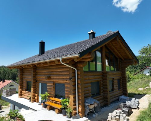 Schrägansicht eines Naturstammhauses mit grauem Satteldach, zwei Kaminen, breiten Dachüberständen und sichtbarer Rundstammkonstruktion