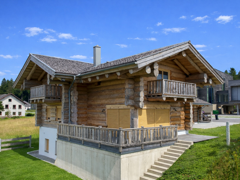 Naturstammhaus mit zwei Holzbalkonen an der Giebelseite, hohem Betonsockelgeschoss, seitlicher Außentreppe aus Betonstufen und großen Fenstern mit gelben Rollläden